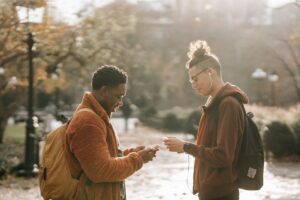Side view of cheerful young friends with backpacks standing in city park and chatting on smartphones on autumn day